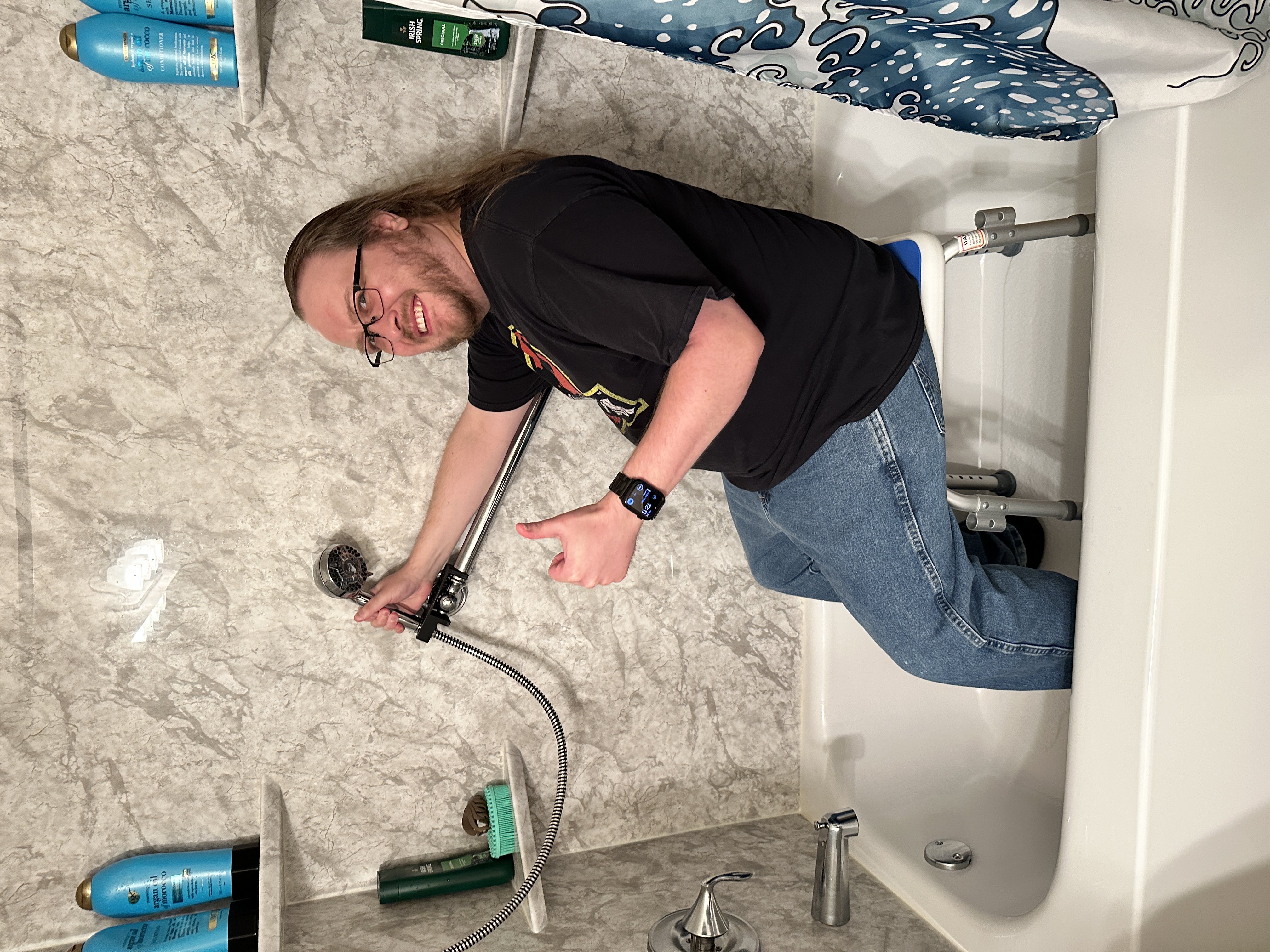 The article author, Tom, sitting on an accessible waterproof shower seat,
fully clothed. He is easily grabbing a shower wand attached to the shower grab
bar with a 3D printed part. He is smiling and giving a “thumbs up” gesture. This
image is illustrating the 3D printed part as a solution to the previous
image.