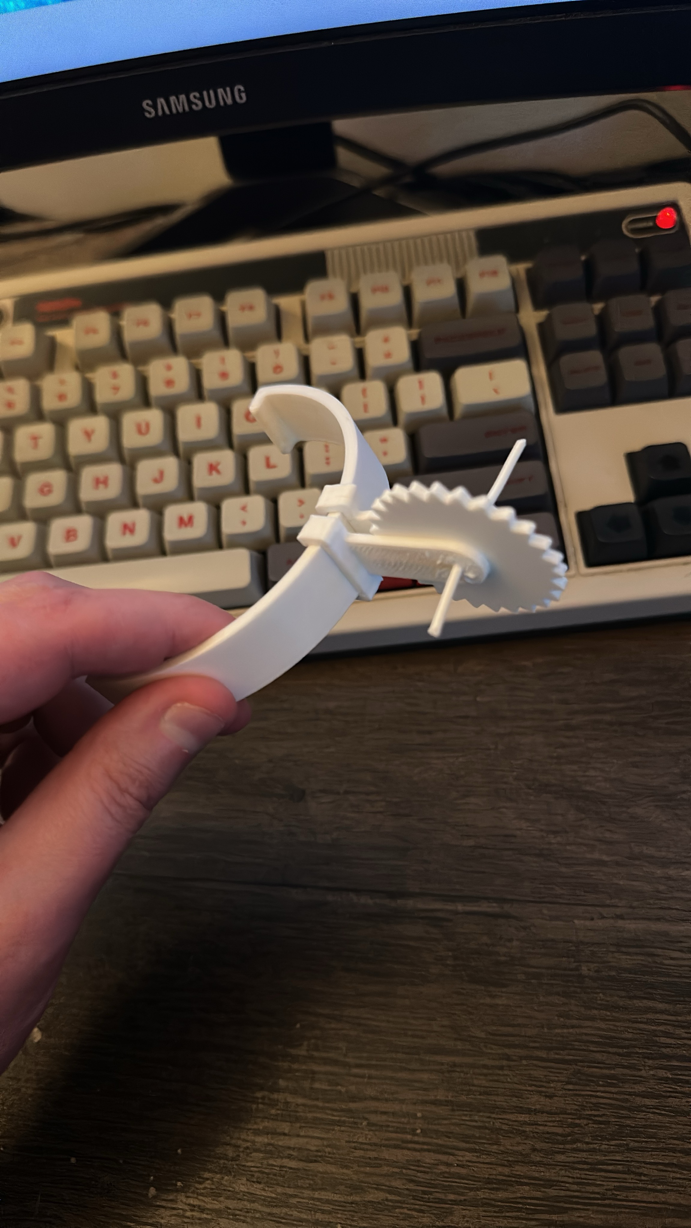 A hand holding a 3D printed Spur and Spur attachment assembly. It is printed
in white PETG. The spur is held aloft by a piece of white PETG filament slotted
between the holes in the SpurConnectors and the hole in the
Spur.